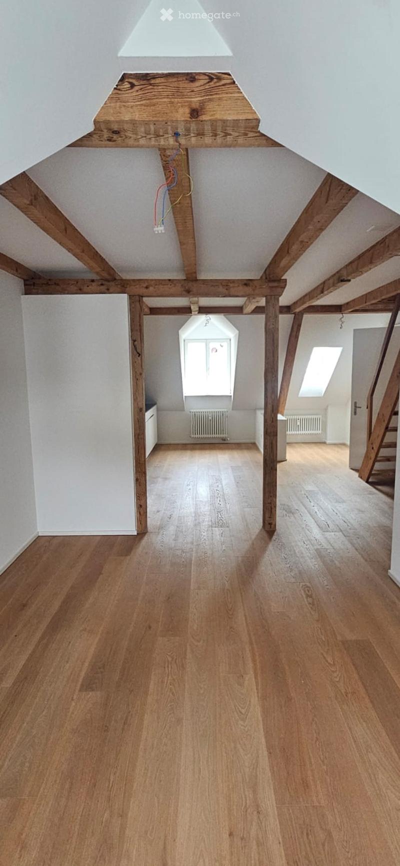 An empty attic space with wooden beams, exposed wooden ceiling, and wooden flooring. The room features skylights and a small window, with no furniture or equipment present.