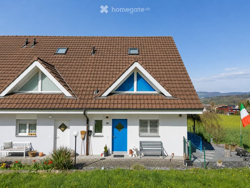 Single-story house with a brown tiled roof, white walls, and blue front door. Features include a small porch with benches, potted plants, and a garden area with a flagpole.