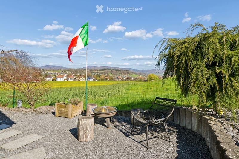 Outdoor area with gravel ground, a fire pit, a chair, and a flagpole with a flag. There are also wooden logs and a planter with plants.