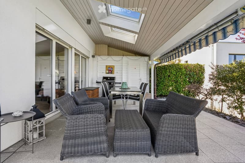 Outdoor terrace with wicker furniture, table, lantern, and grill. Tiled floor and glass doors.