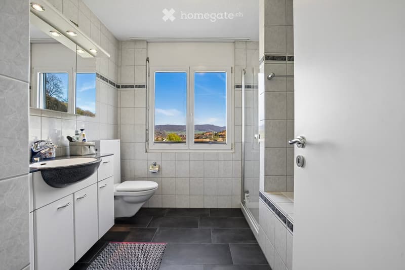 Modern bathroom with white tiles, a sink, toilet, and shower area. Large windows provide natural light.