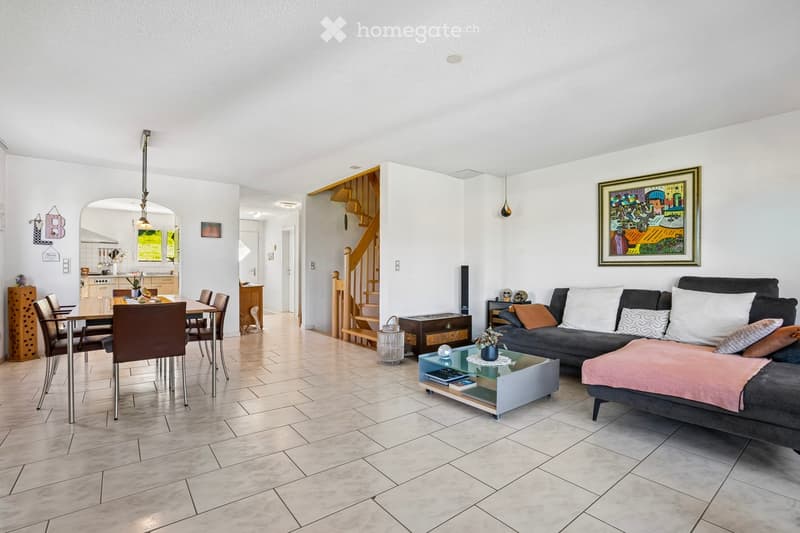 Open-plan living and dining area with a dining table, chairs, and a couch with pillows. A coffee table with a plant and a painting on the wall. A staircase leads to the upper floor.