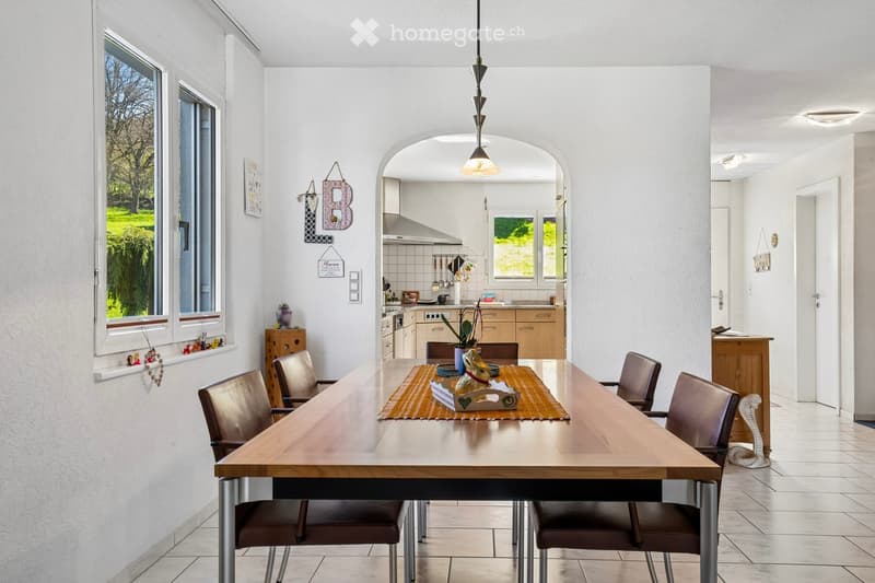 Dining area with a wooden table, leather chairs, and a decorative box on the table. The room has white walls and tiled flooring. A kitchen is visible through an archway.