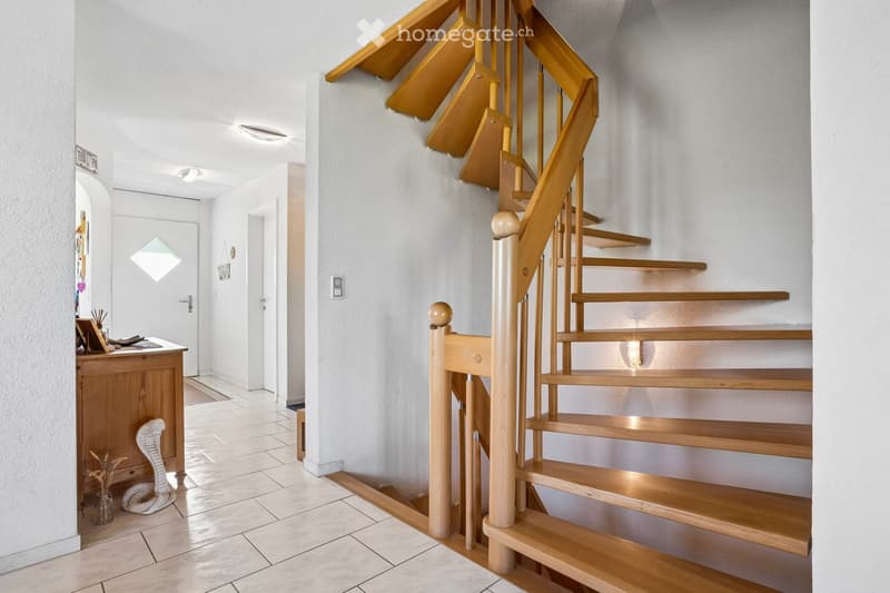 A hallway with a wooden staircase, white walls, and tiled floor. There is a desk with a snake statue and a vase on the left, and a door with a doormat on the right.