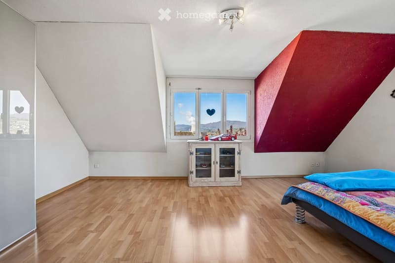 Empty room with wooden floor, white walls, and a red accent wall. A bed with a colorful bedspread and a pillow is placed against the red wall. A small cabinet with a toy car and a lamp on top is near the window.