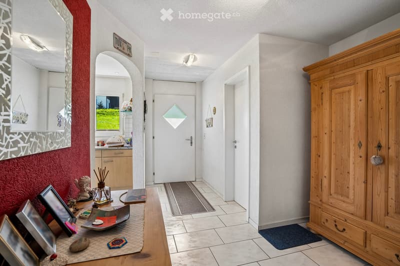 A hallway with a red wall, a mirror, a wooden table with various items, a wooden cabinet, and a wooden wardrobe.