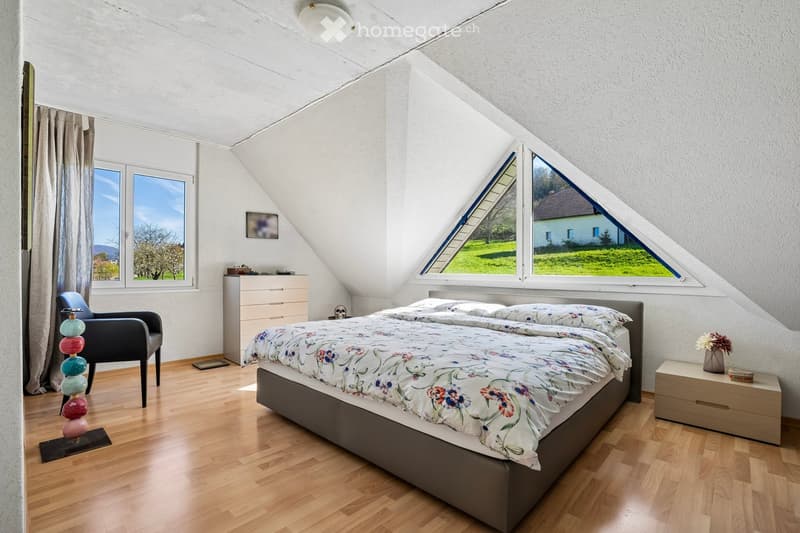 Bedroom with wooden floor, triangular windows, bed, nightstand, dresser, and a chair. The bed has a floral bedspread. The nightstand has a vase with flowers. The dresser has a picture frame on top. The room has a lamp and a rug.