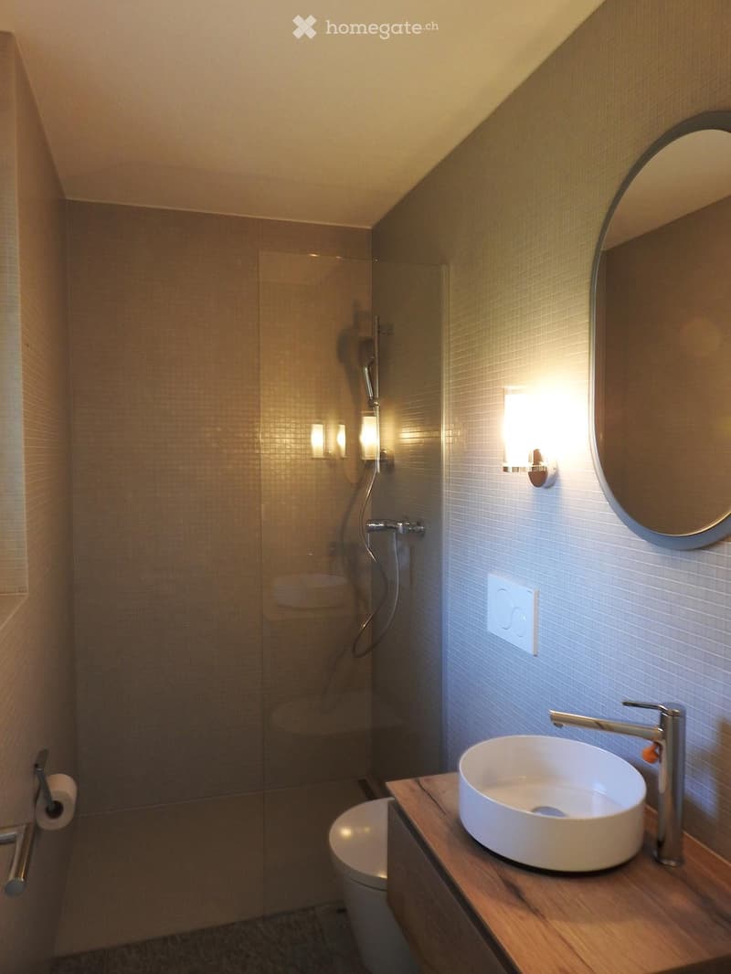 Modern bathroom with a sleek white sink, chrome faucet, round mirror, and a glass-enclosed shower. The space features warm lighting and a wooden vanity.