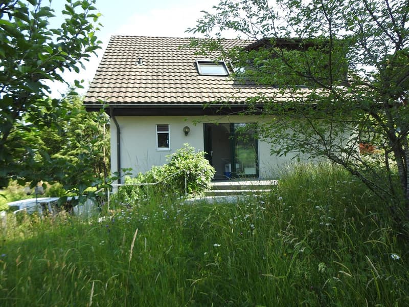 A single-story house with a tiled roof, surrounded by greenery and a parked car in the foreground.