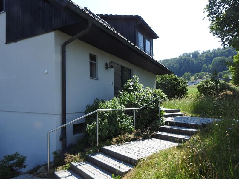 Two-story house with a raised ground floor, featuring a concrete staircase with metal railings leading up to the entrance, surrounded by greenery and a scenic valley view.