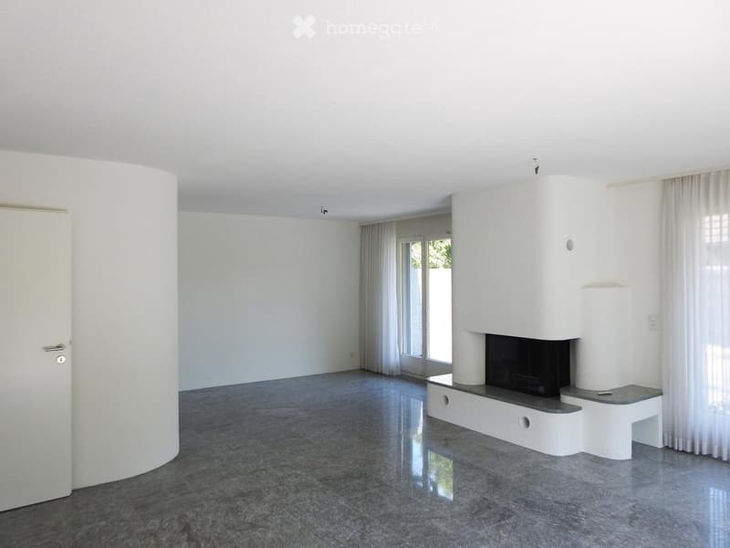 Empty room with white walls, grey tiled floor, built-in fireplace, large windows with white curtains, and a white door.