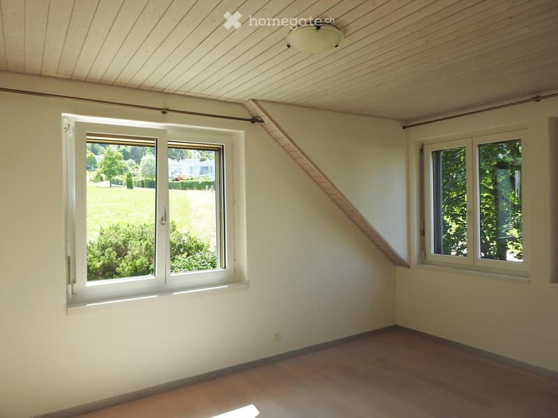 Empty room with wooden floor, white walls, and large windows offering a view of greenery outside. The ceiling has wooden paneling and a light fixture.