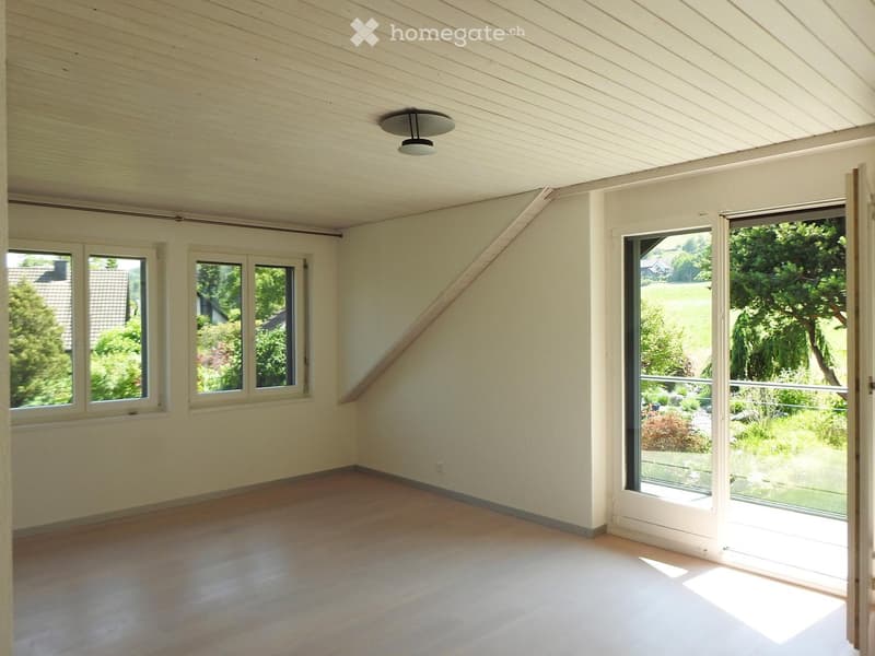 Empty room with wooden flooring, white walls, large windows, and a sliding glass door leading to a garden view.