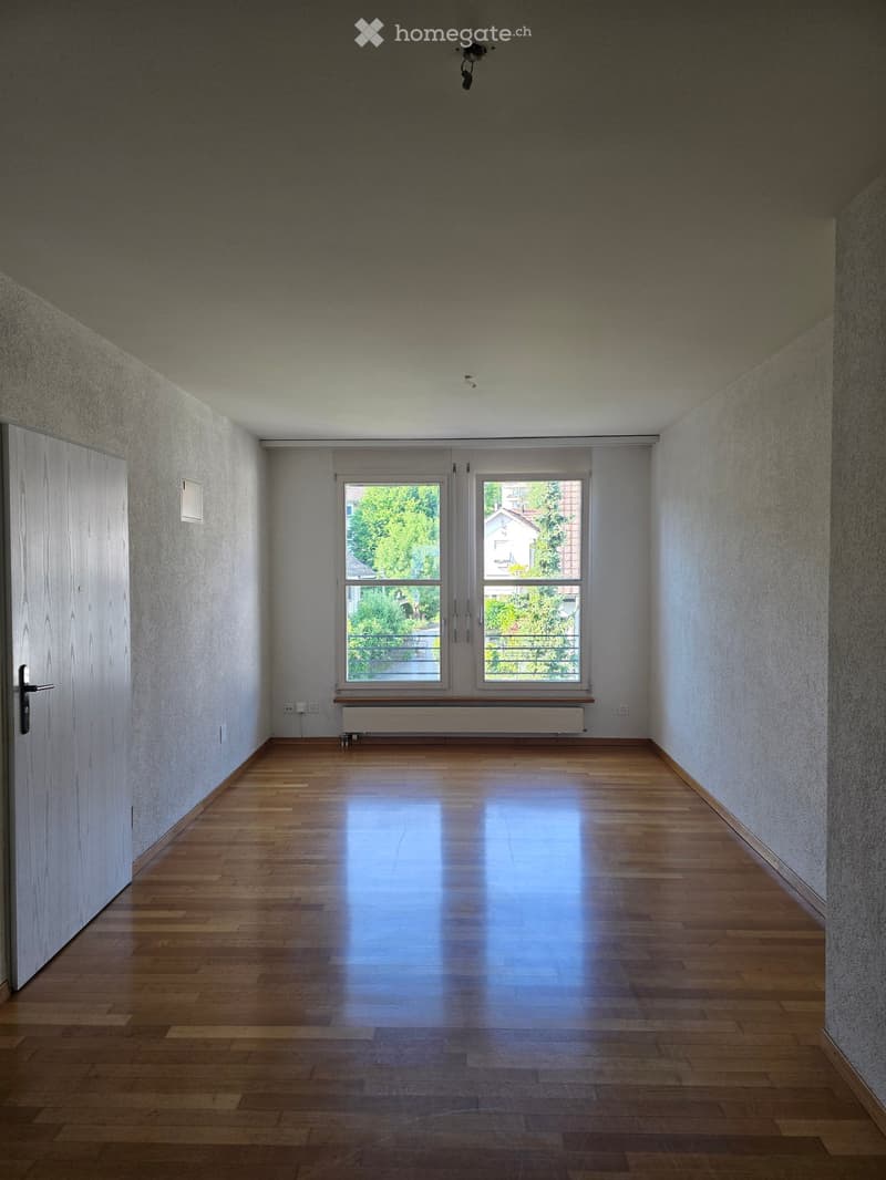 Empty room with wooden flooring, white walls, and large windows with a view of greenery outside. No furniture or equipment is present.