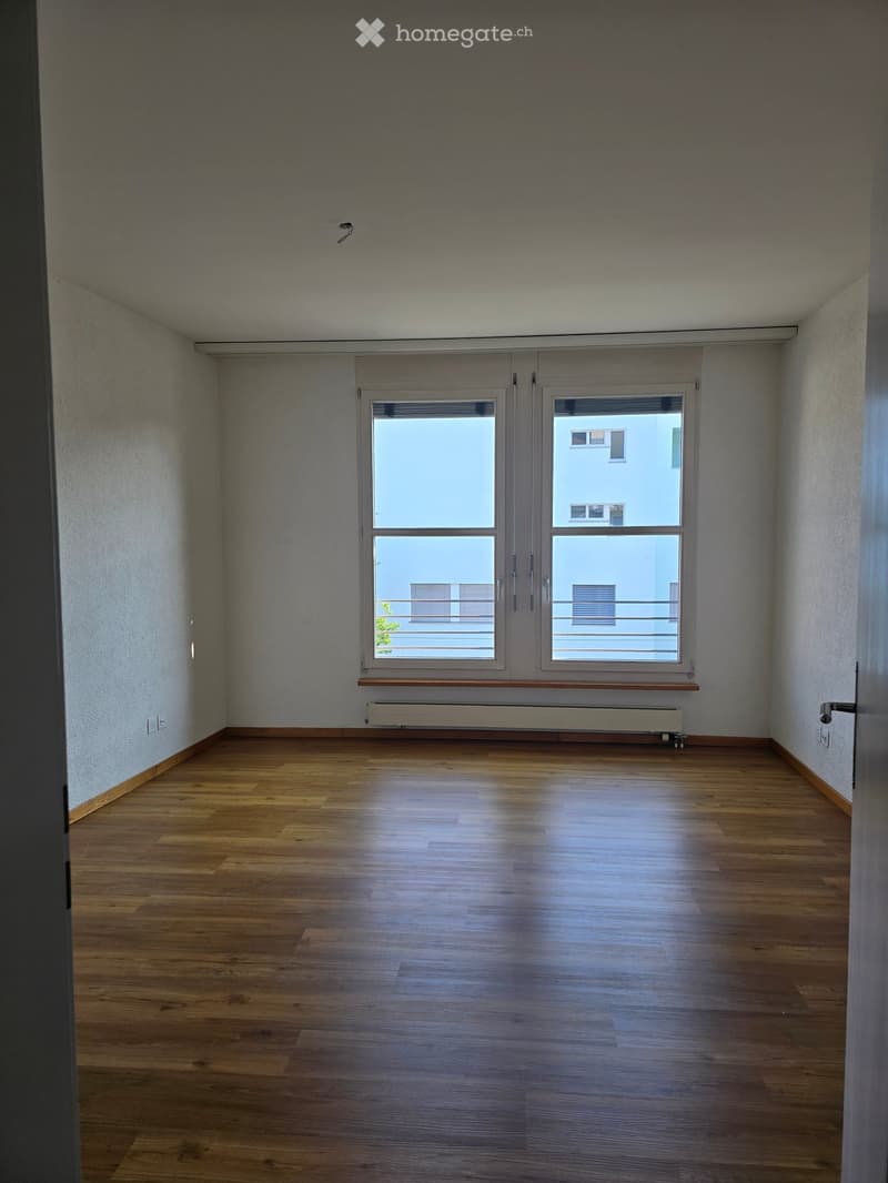 Empty room with wooden flooring, large window with a view of a balcony, radiator, and a door.