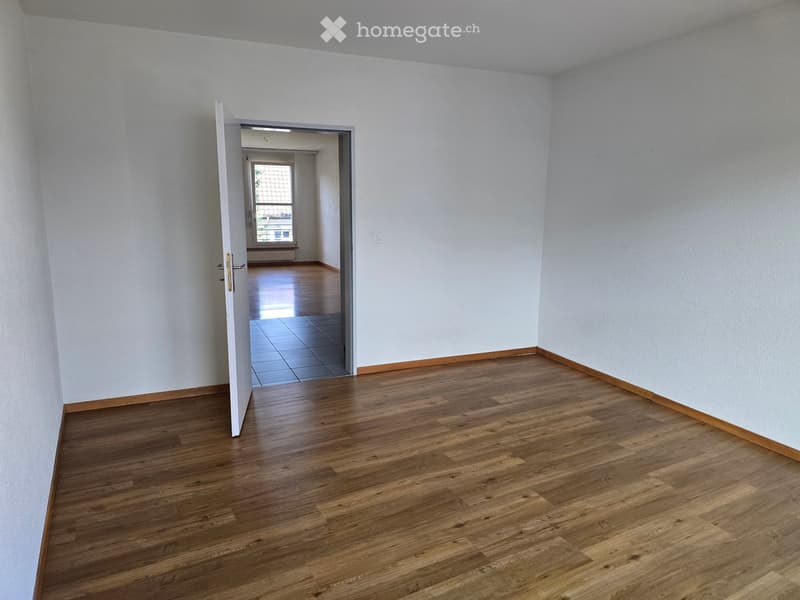 Empty room with light-colored walls and wooden flooring, featuring an open doorway leading to another room with tiled flooring and a window.