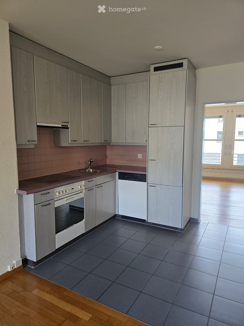 Modern kitchen with grey cabinets, stainless steel appliances, dark tiled floor, and a wooden floor area visible through an open doorway.