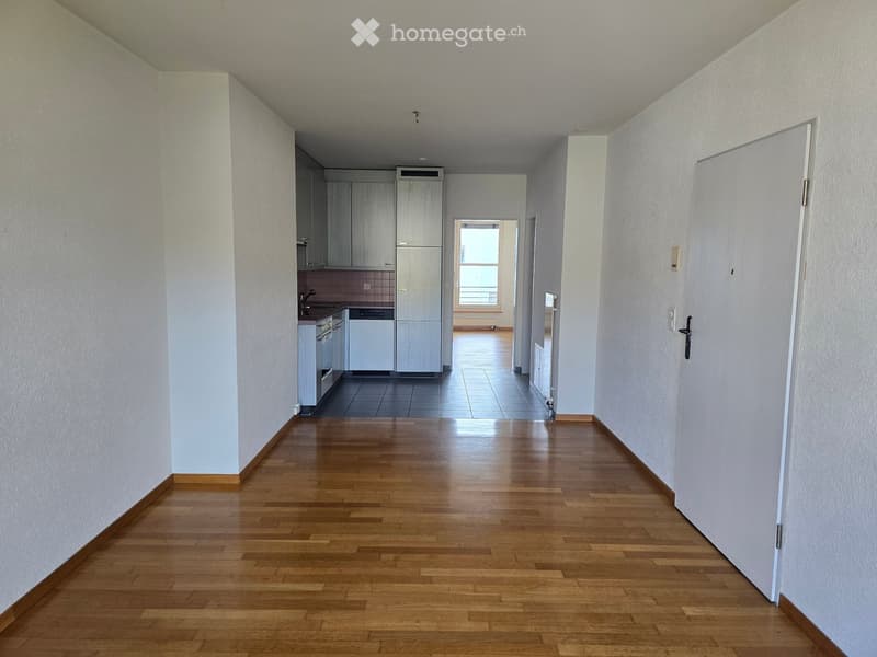 Empty hallway with wooden flooring, white walls, and doors leading to a kitchen area with cabinets and appliances.
