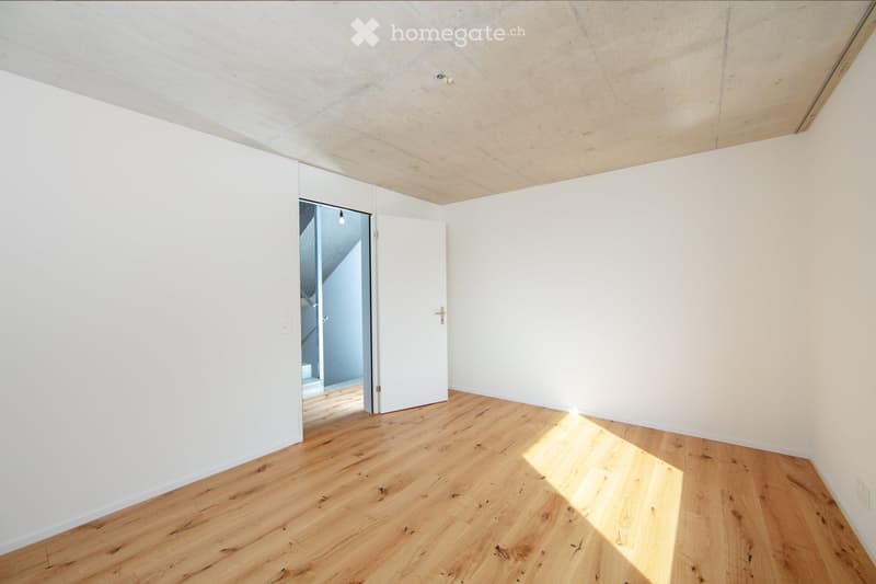 Spacious room with white walls, wooden ceiling, and hardwood flooring. The room has a large window and a door leading to another area.