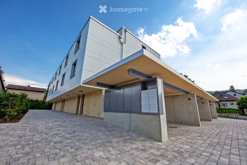 A modern, multi-story building with a flat roof and large windows. The building has a garage or parking area on the ground level, with a paved walkway leading to the entrance. The exterior is primarily composed of light-colored panels and concrete, giving