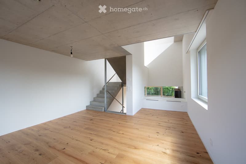 Spacious open-plan living area with concrete ceiling, wooden flooring, and a large window providing natural light. The room features a staircase leading to an upper level.