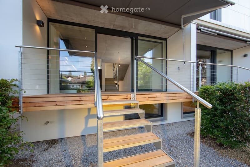 Modern two-story house with concrete exterior, large glass windows, wooden deck with stairs, and metal railing on the balcony.