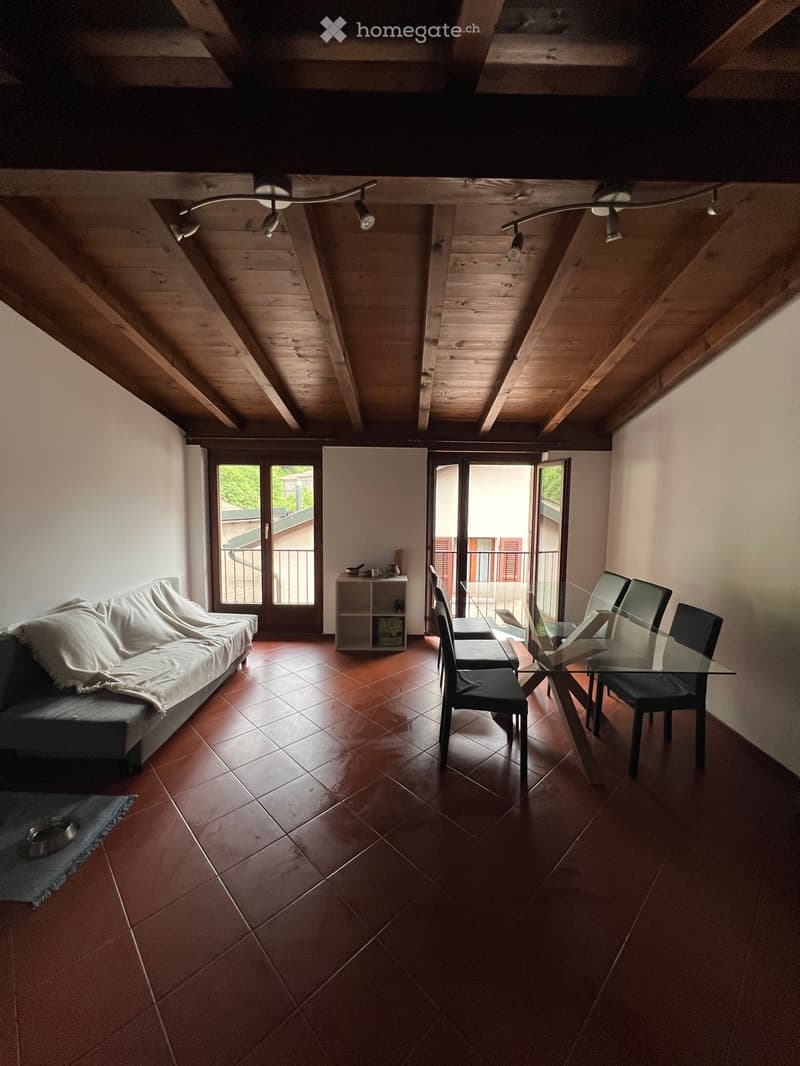 A modern living and dining area with exposed wooden beams, terracotta tiles, and large windows opening to a balcony. Features include a sofa, dining table with chairs, and a small shelf.