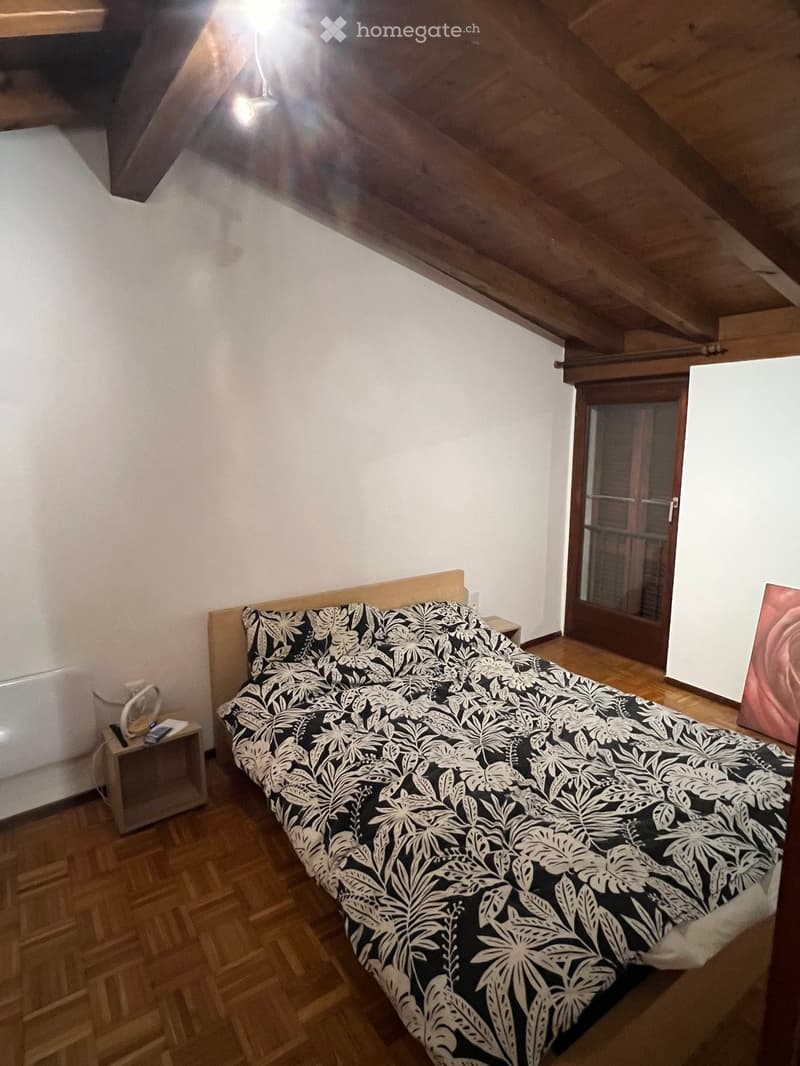 Bedroom with wooden floor, slanted wooden ceiling, bed with black and white patterned duvet, nightstand with fan, and a painting on the wall.