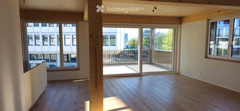 Bright, empty room with wooden flooring, large windows, and a sliding glass door leading to a balcony area.