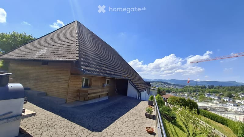 House. Wooden house with a steep roof, large windows, and a spacious patio overlooking a scenic valley with mountains in the background.