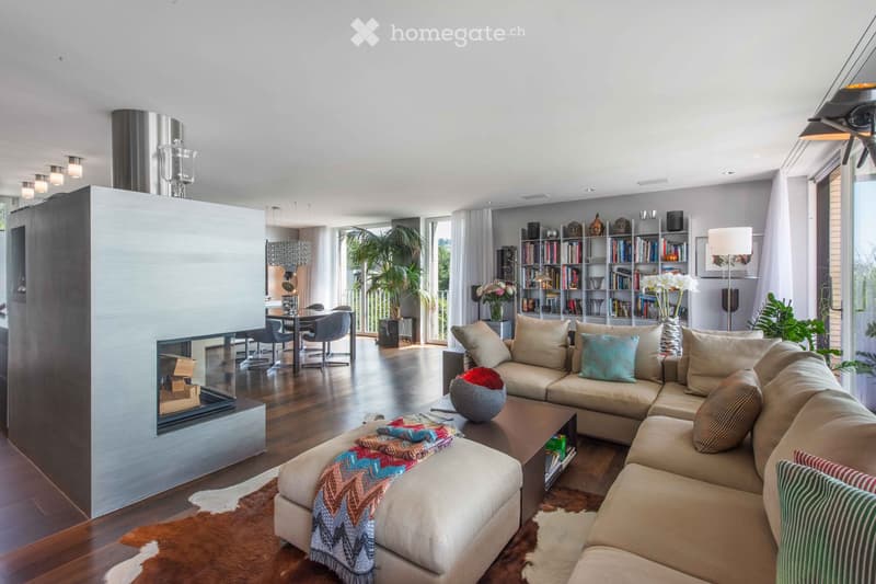 Contemporary living room with fireplace, large windows, bookshelves, glass coffee table, comfy sofa and chairs, area rug, side table, and various decorative items