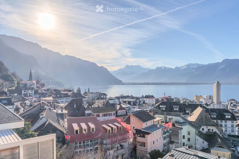 A panoramic view of a town with colorful houses, a lake, and distant mountains under a clear sky.