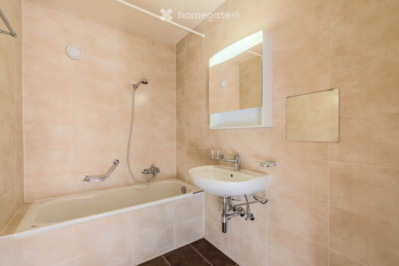 Modern bathroom with beige tiled walls, a bathtub, a sink with a modern faucet, a built-in mirror cabinet with lighting, and a handheld showerhead.