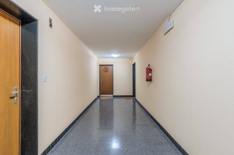 Clean, well-lit hallway with light-colored walls, dark tiled floor, and wooden doors. Features a fire extinguisher mounted on the wall and a small decorative wreath on one door.
