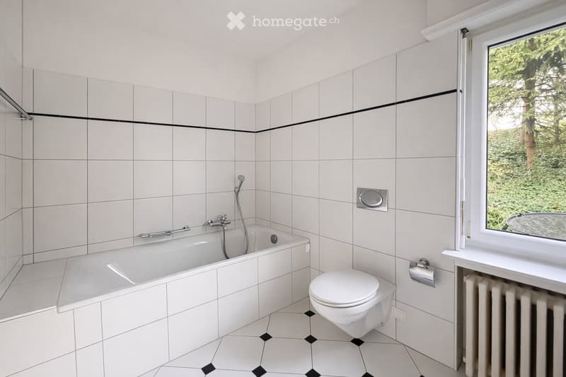 Modern bathroom with white tiles, a bathtub, toilet, sink, and radiator. Features a large window with a view of greenery.