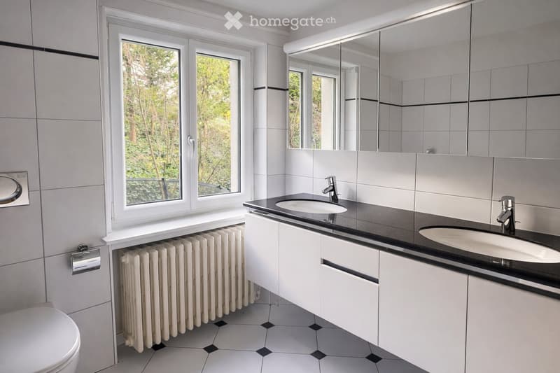 Modern bathroom with white tiles, black countertop, double sink, large mirror, toilet, and radiator under window.