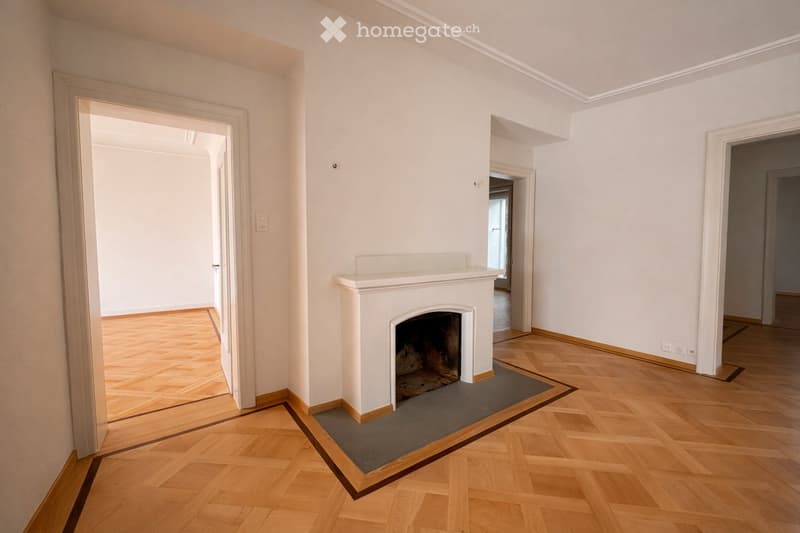 Empty room with wooden flooring, white walls, and a fireplace. The room has multiple doorways leading to other spaces.