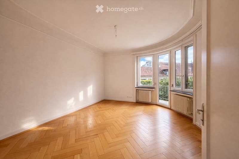 Empty room with wooden herringbone flooring, white walls, large windows with view, radiators, and a door.