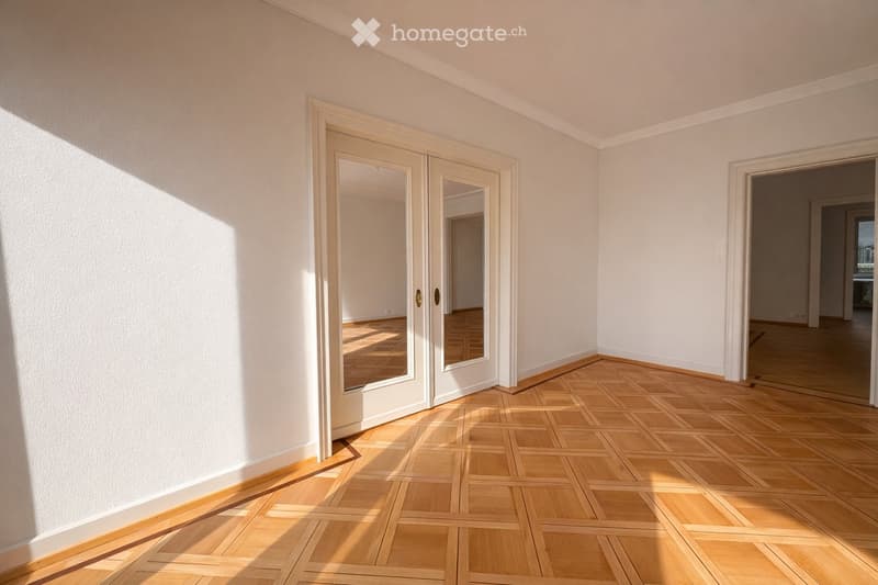 Empty room with light wooden flooring and white walls, featuring a large mirrored double door and an open doorway leading to another room.