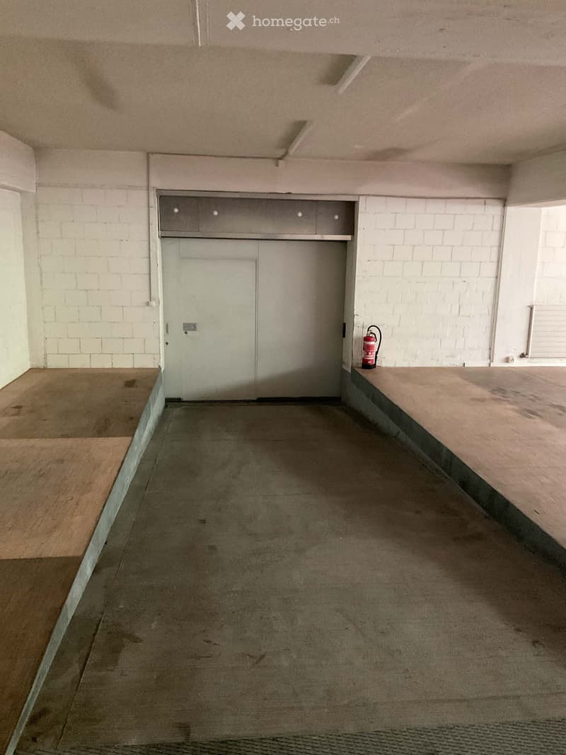 Unfurnished Room. Empty room with concrete floor, white brick walls, and a closed garage door. A fire extinguisher is placed on a wooden platform to the right.