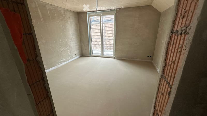 Empty room with unfinished walls and floor, featuring a large window with a view of a tiled roof outside.
