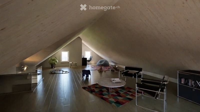 Loft-style attic room with wooden floors, slanted ceilings, and large windows. Features a round coffee table, chairs, a small cabinet, a plant, and a colorful rug.