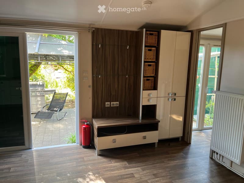 A modern, well-lit room with wooden flooring, white cabinets, wicker baskets on a shelf, a radiator, a fire extinguisher, and a comfortable chair near the sliding glass door leading to an outdoor area.