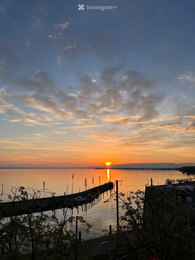 Garden / Outdoor Area. View of a serene lake at sunset, with a dock extending into the water and distant mountains visible under a colorful sky.