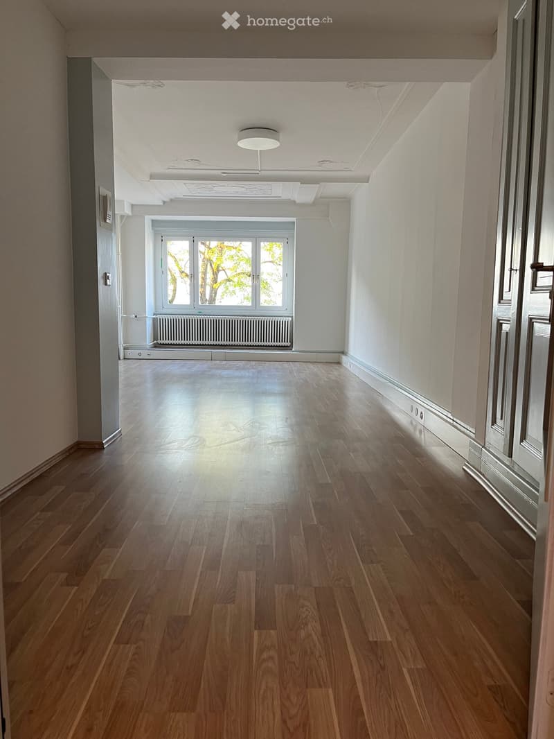 Corridor / Hallway. Empty hallway with wooden flooring, white walls, and a large window at the end allowing natural light. The ceiling features recessed lighting and a visible air conditioning unit.
