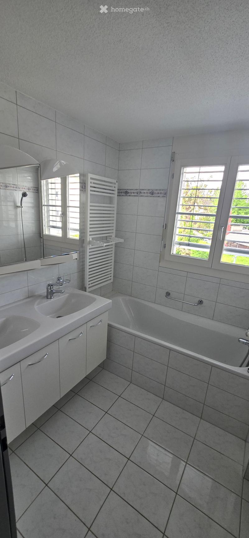 Bathroom. Modern bathroom with a white sink, bathtub, mirror, and radiator. The room features tiled walls and floor with large windows providing natural light.