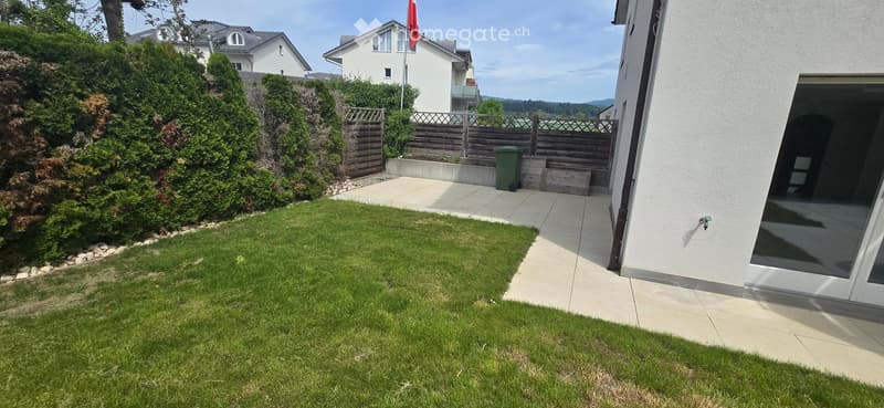 Garden / Outdoor Area. Well-maintained garden with a neatly trimmed lawn, a paved patio area, and surrounding greenery, including shrubs and trees. A wooden fence encloses the garden, and a green trash bin is visible near the fence.
