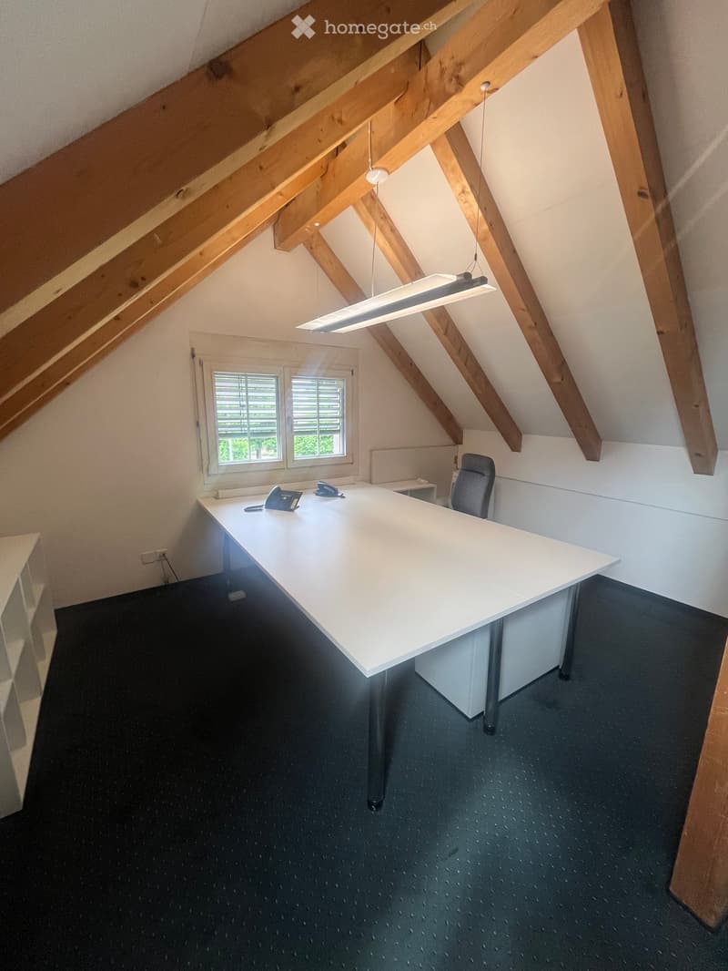 Office space with exposed wooden beams, a large white desk, a black office chair, and a telephone on the desk. The room has a slanted ceiling and a window with blinds.