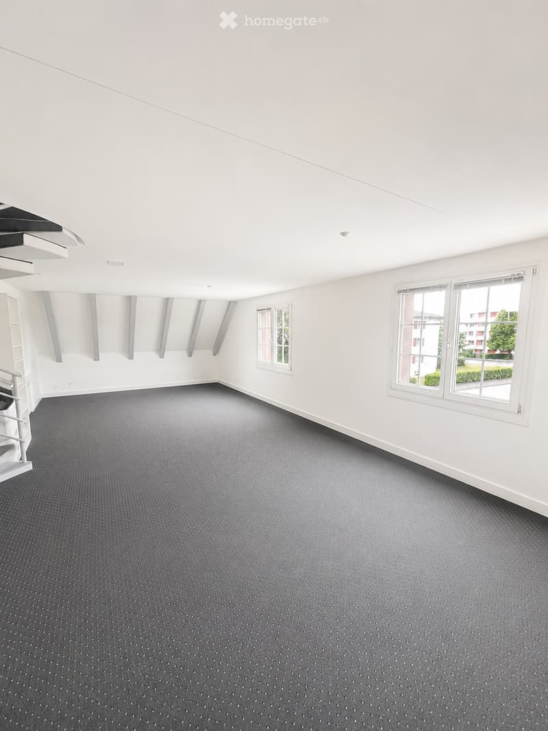Empty room with white walls, grey carpeted floor, and multiple windows allowing natural light. No furniture or equipment present.
