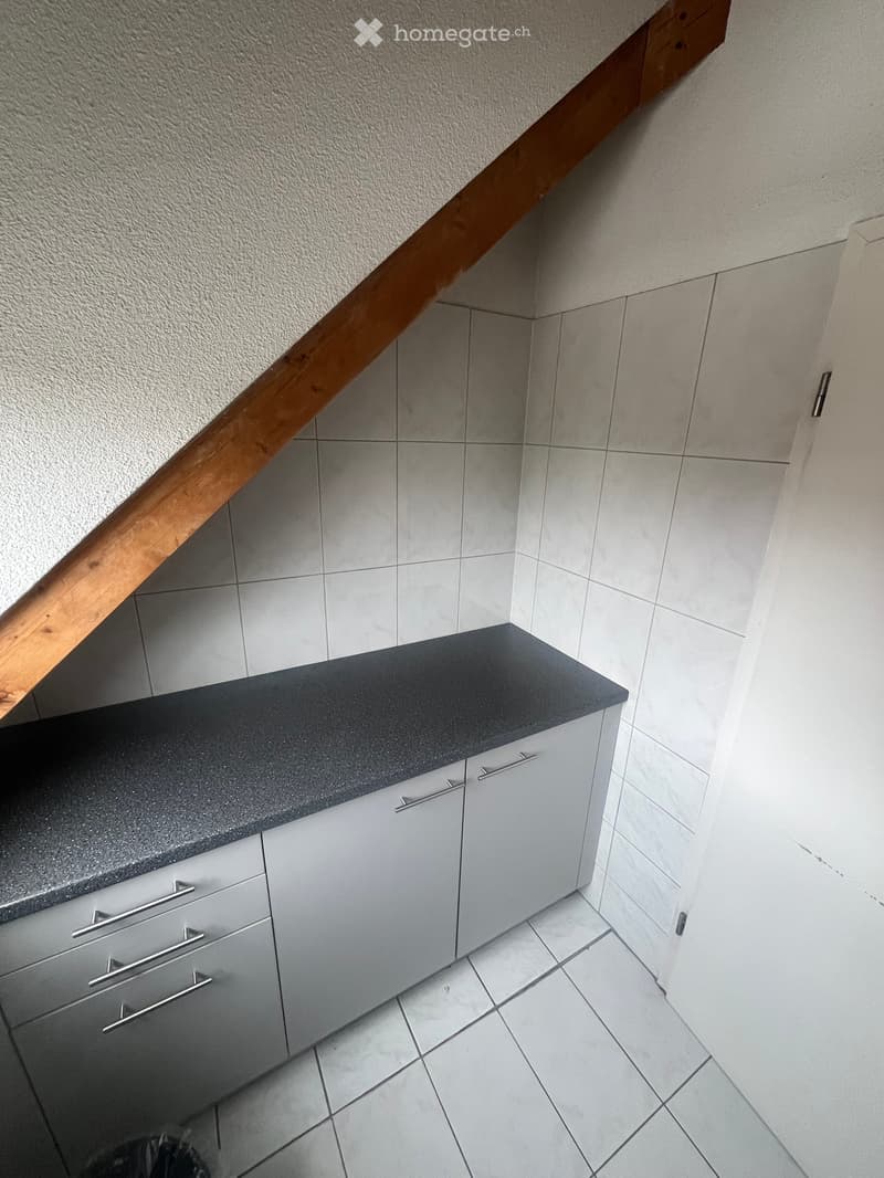 Modern kitchen with grey cabinets, black countertop, and tiled floor. Wooden staircase visible in the background.